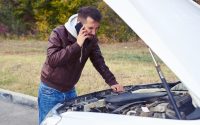 discontented man talking on the phone and looking under the hood of breakdown car at outdoor