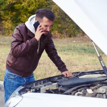 discontented man talking on the phone and looking under the hood of breakdown car at outdoor